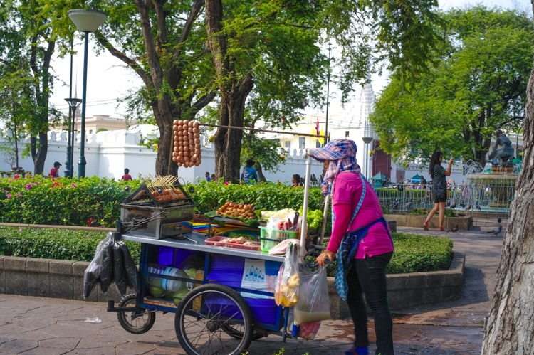 A woman wearing bright pink selling all types of street food from a cart at a temple in Bangkok.